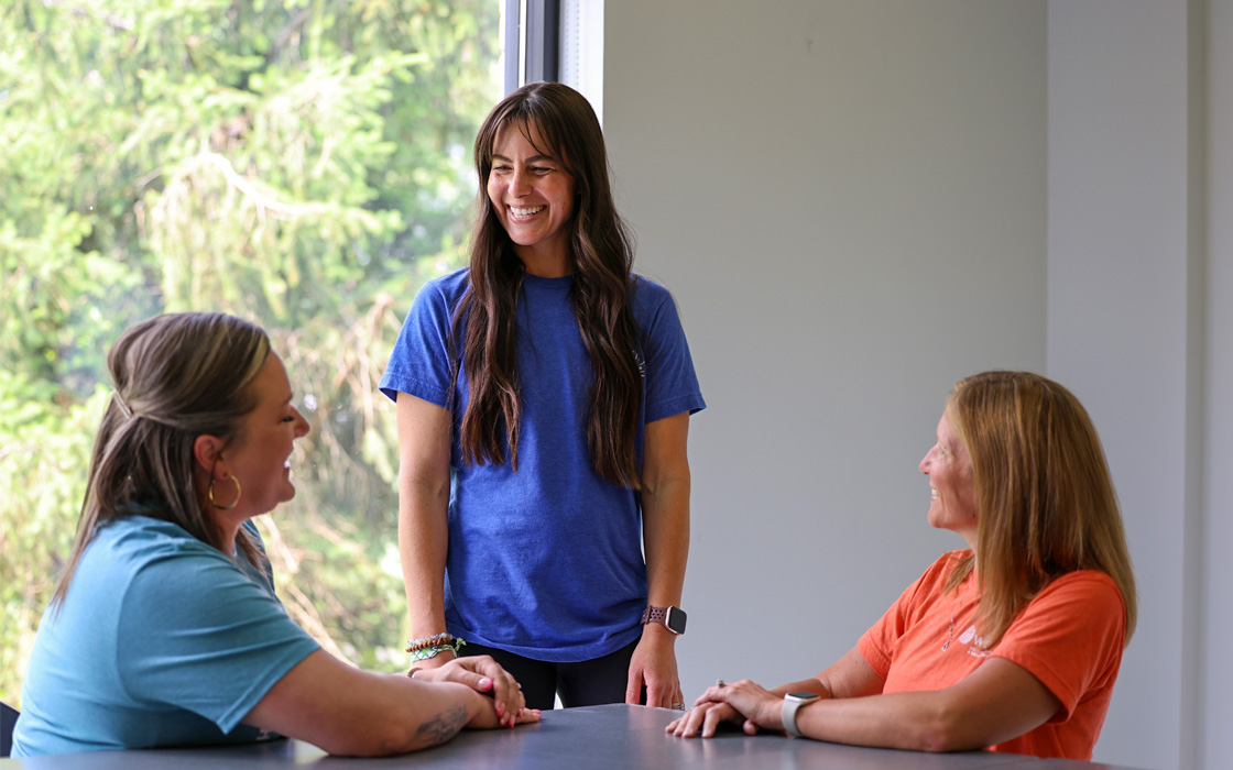 Photo of two women sitting at a table talking to another standing up